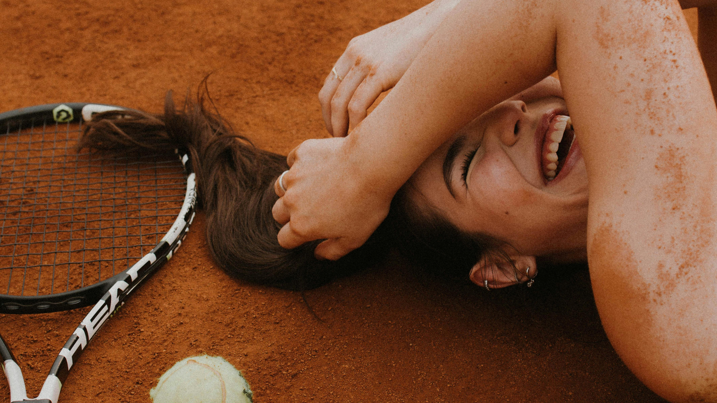 Woman laughing and laying on the ground next to a tennis ball and racket.